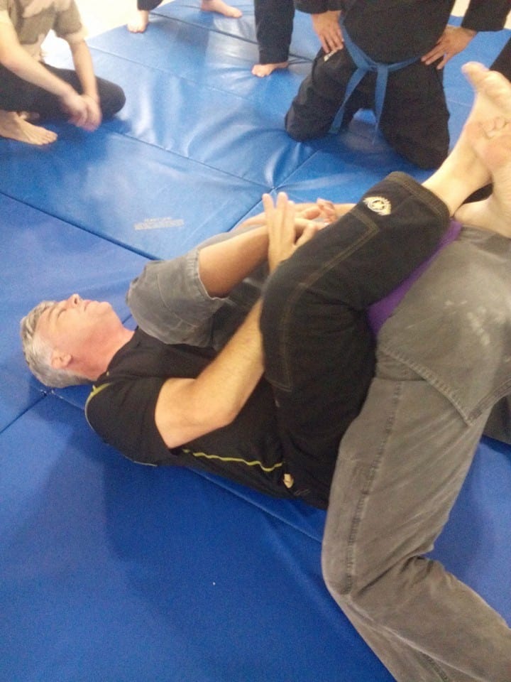 Adult male instructor demonstrating a self-defense technique on a student during a martial arts training session on blue mats, with other participants observing.