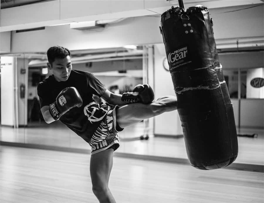 Young martial artist delivering a powerful kick to a heavy bag in a training studio, emphasizing fitness and power development in martial arts.